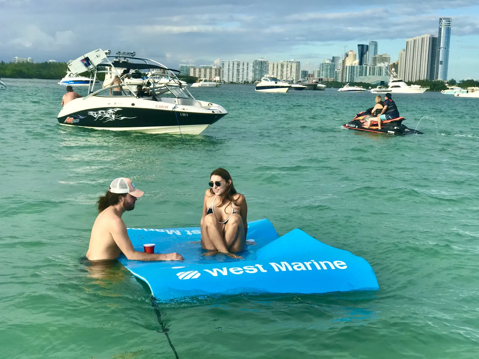 People relaxing in shallow turquoise water at a sandbar with boats, a jet ski and the Sunny Isles skyline behind them